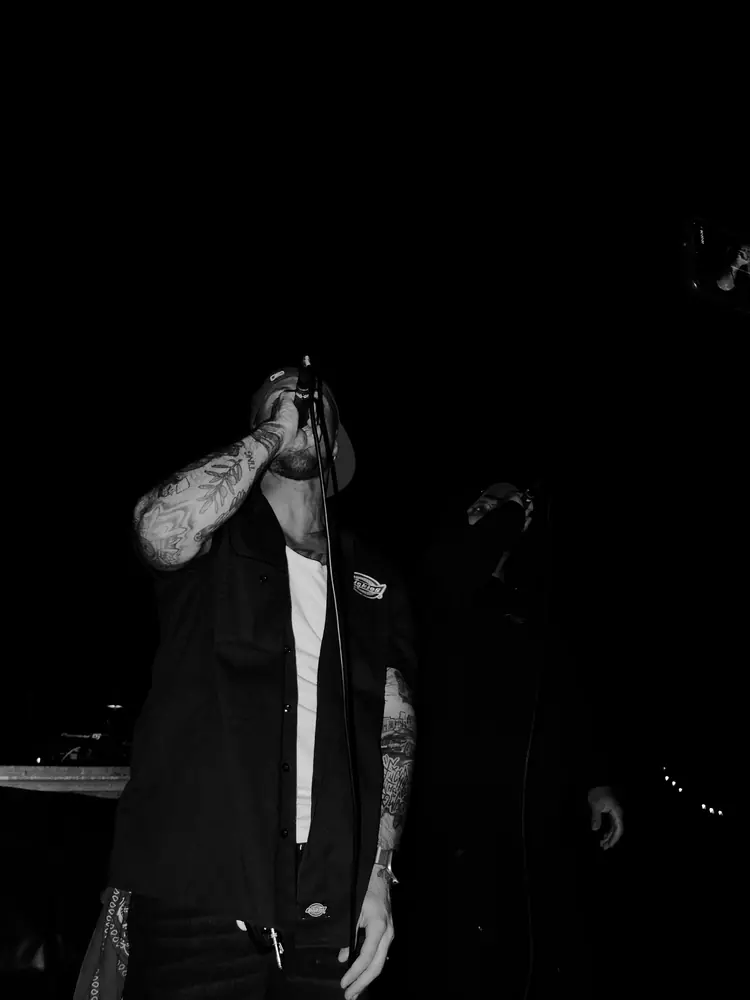 Foto in bianco e nero di un live: un MOFO rappa al microfono, braccio tatuato, sul palco al buio
